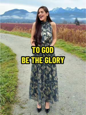 A woman is standing on a paved path with a mountain range in the background. She is dressed in a long dress with a floral pattern and is smiling widely. The text overlay, 'TO GOD BE THE GLORY,' is prominently displayed in a yellow box.