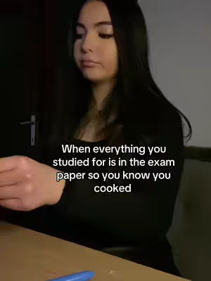 A young woman with long dark hair looks off to the side with a slight smile. Text overlay reads, "When everything you studied for is in the exam paper so you know you cooked." She is seated at a desk, and a pen is visible near the bottom. The background is dimly lit.