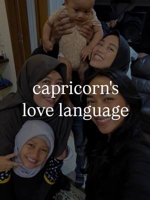A candid photo of a multi-generational group, likely indoors. The main subject group incorporates a smiling child held by a person facing the camera, four other people, including two wearing hijabs, are clustered closely together. The background is blurry, possibly a room or apartment setting, with warm lighting.