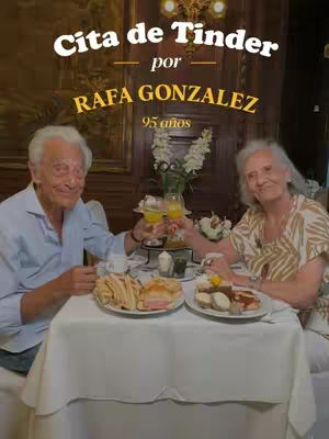 An older couple is seated at a table set for a meal. They are dressed casually, with the man in a light blue shirt and the woman in a patterned blouse. The table is covered with a white tablecloth, and plates of food are visible along with glasses. The background is dark, but appears to be a restaurant with wooden accents. Text overlays at the top and in the center provide context.