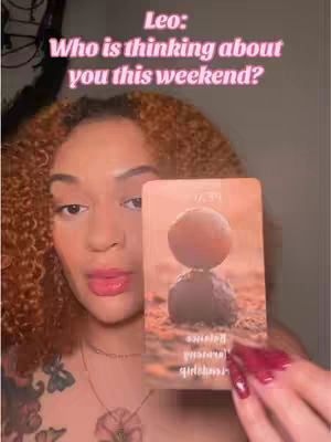 The cover image shows a woman holding a tarot card. The card features an image of two stones with a circle balancing on top. Text overlay reads, 'Leo: Who is thinking about you this weekend?' The woman has curly brown hair and is wearing makeup. Her red nails are prominent.