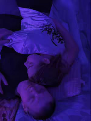 An overhead shot of two people lying in bed, embracing. The scene is lit with purple light, creating a dim, intimate setting.
