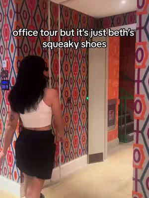 A woman is walking down a hallway, seen from the back. She is wearing a white crop top and a black skirt. The hallway has wallpaper with a repeating geometric pattern in red, orange, and black, the lighting is from an overhead source.