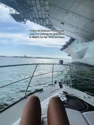 A shot is taken from a moving boat, looking forward. The viewer's legs are in the foreground, with a boat railing and the water in the middle ground. In the background, an enormous ship rests in the water.
