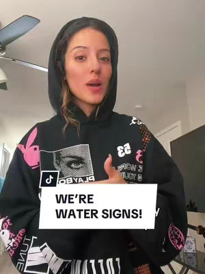 A woman wearing a black hoodie with a hood up, looking directly at the camera with a neutral expression. A white rectangle with the text "WE'RE WATER SIGNS!" is overlaid at the bottom. The background is a well-lit living room.