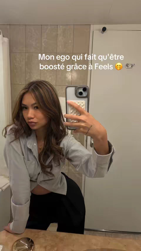 A woman takes a selfie in a bathroom. The background features a patterned tile wall. She is wearing a grey shirt and black pants and holding a phone up, likely in the act of taking the photo.