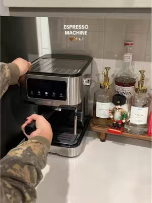 A person is holding an espresso machine, with a clean countertop and various bottles and a small figurine in the background. The image has a natural lighting, and the focus is on the espresso machine.
