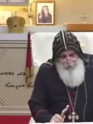 A man in religious attire is seated, facing the camera. He has a white beard and is wearing a black robe with a dark hood that has a patterned design. On his chest is a cross pendant. Behind him are several objects, including a gold head covering. A framed image of a woman hangs on the wall, and there is also what appears to be writing on the wall behind him.