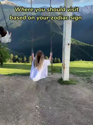 A woman is swinging on a swing set, positioned with green mountains and trees in the background. Text overlaid says 'Where you should visit based on your zodiac sign.' Another person is on a swing to the left.