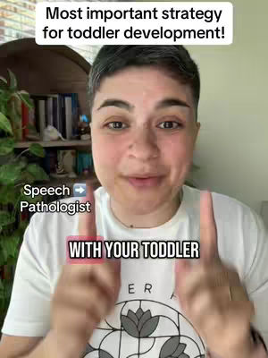 A person with short dark hair and glasses points towards the camera with two fingers. A text overlay in a white rectangle says, "Most important strategy for toddler development!" and another text element says "Speech Pathologist". The background is slightly blurred with a hint of a bookshelf and a vibrant green plant. The person is wearing a white t-shirt with a graphic at the bottom.