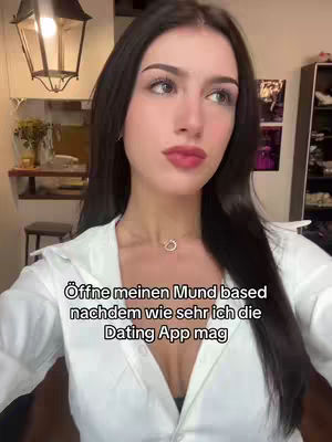 A woman with long dark hair is shown from the chest up, looking towards the upper right. She wears a low cut white shirt with a necklace. The background shows a room with overhead lighting.