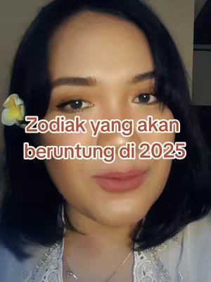 A close-up shot of a woman's face. She has dark hair, styled away from her face, and her eyes are looking forward. A white and yellow flower is tucked behind her left ear. The image has text overlay that reads 'Zodiak yang akan beruntung di 2025'. The woman is wearing makeup, including eyeshadow and lipstick.