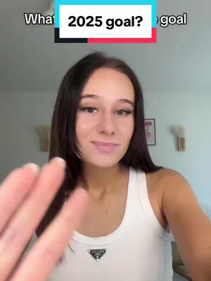 A young woman with long dark hair and a white tank top smiles at the camera. The overlaid hook text is in a colored rectangle with a question about a 2025 goal. She is partially obscuring the frame with her outstretched hand. The background is a lightly colored wall with a bright light source. The overall composition is bright and inviting.
