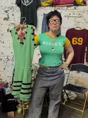 A woman with short dark hair and glasses stands indoors, holding a green dress on a hanger. She's wearing a green shirt and gray pants, posing in front of a wall with various clothing items hanging in the background.