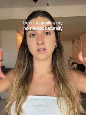A woman with long, light brown hair is centered in the frame, looking directly at the camera. The background is a softly blurred room interior. Text overlayed on top states "how I'm healing my hormones naturally".