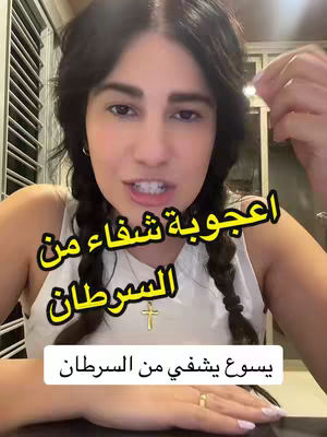 A woman with dark hair in braids is centered, looking at the camera and speaking. Above her, text in Arabic reads " اعجوبة شفاء من السرطان ", meaning a miracle of healing from cancer in English. Below that, text in Arabic says "يسوع يشفي من السرطان", which means "Jesus heals from cancer". The background is a room with a blurred view, and she is wearing a white top and a cross necklace. There is a gold ring on one of her fingers. Overlaying elements of the text create a layered effect.