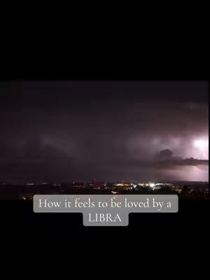 A dark, vertical image with a night sky filled with dark clouds and lightning strikes. Below, a city skyline is visible, with lights illuminating the buildings. A rectangular text overlay in a light gray box reads: “How it feels to be loved by a LIBRA.”