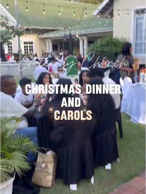 An outdoor scene at a Christmas dinner, featuring people seated at tables decorated with black tablecloths. The photo is shot at a slight downward angle, capturing the event's ambiance. Overhead, string lights hang. In the background, buildings and greenery add depth to the scene. Text overlays read 'Christmas Dinner And Carols' in bold on top of the people at the table, and the caption includes mentions of 'Gateway Church,' indicating a church gathering.