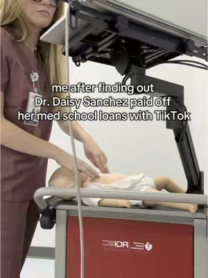A woman in glasses wearing a maroon scrubs shirt, is standing, looking down, and pointing at the chest of a baby mannequin lying on a hospital bed.