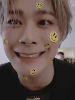 A close-up shot of a young man with fair skin is shown smiling with a wide-open mouth, partially revealing his braces. Yellow smiley face stickers are placed on his face and neck. The background is softly blurred, with hints of a room visible.