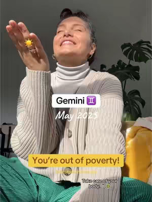 A woman with her right hand raised, displaying a ring with a yellow sun design. She is smiling towards the right, with her eyes closed. She is wearing a white turtleneck under a beige, ribbed cardigan. The background consists of a plant and soft, natural light. Text overlays appear in a white rectangular box reading "Gemini ♊️
May 2023" and another yellow box with the text "You're out of poverty!".