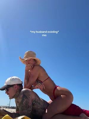 A sunny outdoor shot with a clear blue sky as the backdrop. In the foreground, a person is sitting on the other person's lap with their head resting on the other person's chest. Text that says '*my husband existing* me:' is overlaid on the sky.