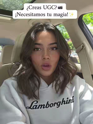 A young woman is seated in a car, facing the camera with an open mouth expression. She wears a white hoodie with the word 'Lamborghini' printed on it. The background shows the car's interior, including the seats, windows, and an overhead view of the sky. Text overlays, in a white rectangle, ask '¿Creas UGC?', with a camera icon, and state '¡Necesitamos tu magia!'.
