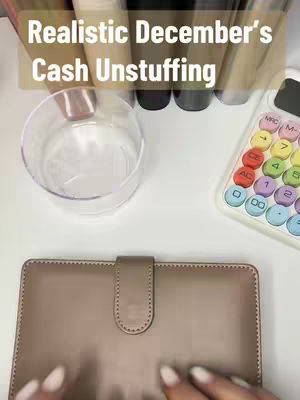 A top-down shot showcasing a desk setup. The image is dominated by neutral tones and soft lighting. A beige leather wallet-style binder rests at the bottom center, subtly reflecting the light. To the left, a clear glass bowl is visible, while on the right, a calculator with colorful buttons adds a playful contrast. Behind these items, a collection of cylindrical containers, possibly for makeup or storage, are positioned in a row. The overall composition suggests organization, budgeting, and potentially, a makeup-related activity, all framed from a high-angle perspective.