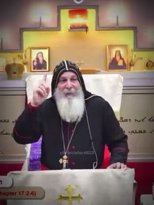 A man with a long white beard and a black and white ecclesiastical hood is standing at a podium. He is raising his right hand with his index finger pointing up. In the background, there is a decorated area with portraits and a golden crown. The background is a light brown with columns. A desk with a cross. Below the host is another cross.