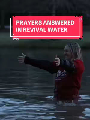 A woman stands waist-deep in water, arms outstretched to the sides, appearing to be in a baptism or similar religious ceremony outdoors. A red rectangular banner with white text is superimposed on the image, indicating "Prayers Answered in Revival Water". The scene is overcast with soft lighting.