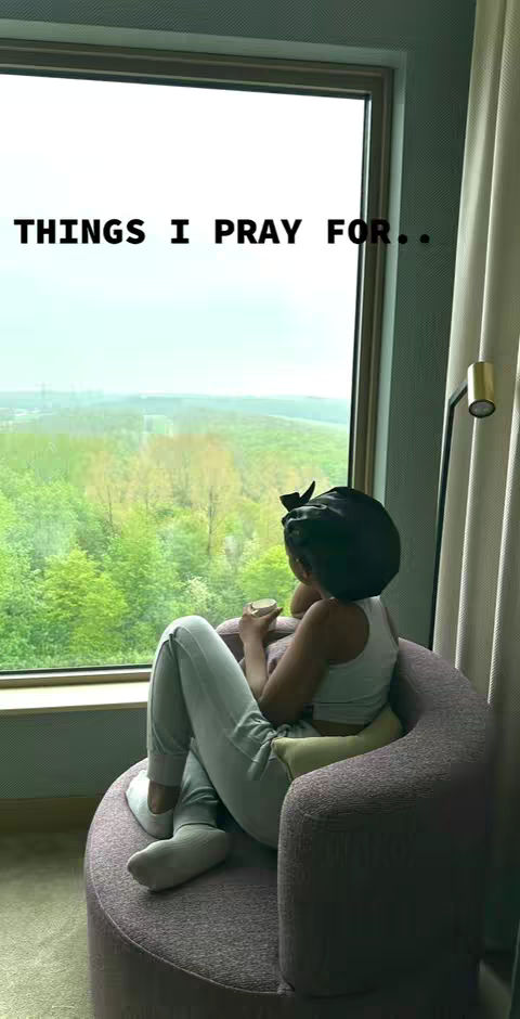 A woman is seated in a round, mauve armchair, gazing out a large window at a verdant, tree-filled landscape. The text "THINGS I PRAY FOR.." appears at the top. The scene is peaceful, with soft lighting and a focus on introspection.
