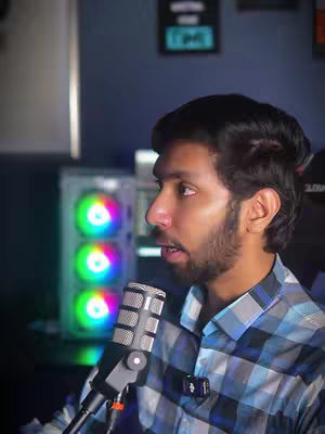 A man with dark hair and a beard is speaking into a microphone. He is wearing a plaid shirt. Behind him is what appears to be a computer setup, including a computer case with RGB lights.