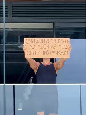 A person stands outdoors holding up a handmade cardboard sign that reads: "CHECK IN ON YOURSELF AS MUCH AS YOU CHECK INSTAGRAM."