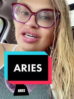 A close-up shot of a blonde woman wearing large, red-rimmed glasses. She has full lips and is speaking. Overlayed on the image is a black box with the word 'ARIES' in white text, and a smaller banner below with the same word. The background is blurred, indicating a car interior.