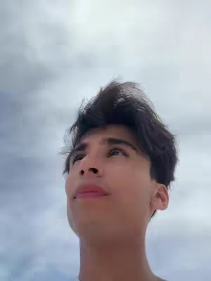 A young man looks up at the sky with an expression of hope or wonder. His dark hair is styled upward, and the background is a blurry gradient of light and dark grey, suggesting a cloudy sky.