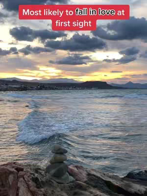 A scenic image of a beach at sunset. The sky is filled with colorful clouds with the sun peeking through gold and orange hues. The ocean water reflects colors of the sky, with waves gently rolling to the shore. A pile of rocks sits in the foreground.
