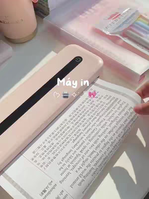 The image shows a close-up view of a desk setup. A light pink printer is prominently displayed in the center, with a black stripe. A book with text in multiple languages is placed partially underneath the printer. Other study supplies like colorful pens and a pink cup are arranged on the desk, contributing to a soft, organized aesthetic.