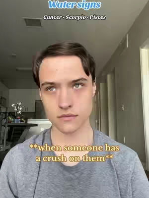 A selfie style shot featuring a young man with short brown hair and a light gray t-shirt. Overlayed at the top of the frame are text lines reading 'Water signs' in a baby blue font, followed by zodiac signs 'Cancer-Scorpio-Pisces'. Overlayed at the bottom of the frame is the text '**when someone has a crush on them**' in a light yellow font. The man is positioned centrally, staring towards the camera with a neutral expression. The background includes interior elements such as doors, walls, and ceiling. The framing is from the chest up.