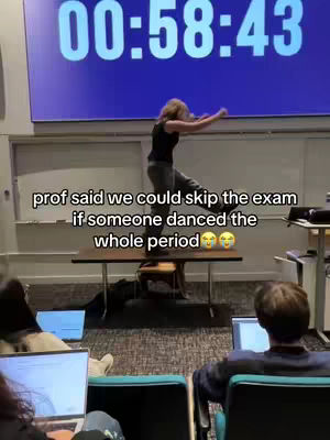 A woman dances on a table in a classroom, students are sitting with laptops. A digital timer is on the screen in the background.