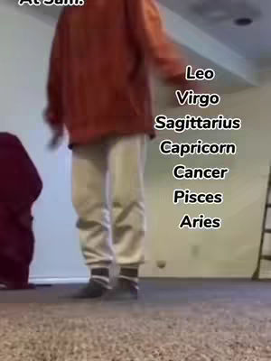 A person in an orange hoodie and light pants stands in a room, partially visible. To the right, a list of zodiac signs is displayed vertically in white text against the wall.