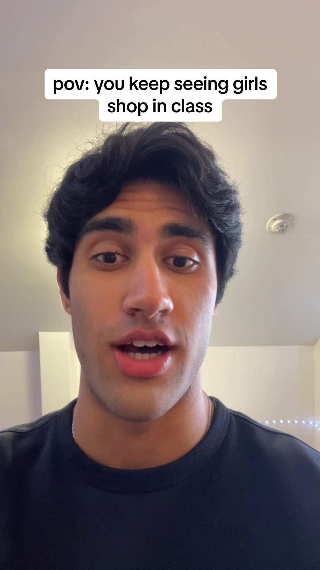 A selfie-style video cover image features a young man with dark hair, looking directly at the camera with a conversational expression. Above him, white text on a black rectangle reads, "pov: you keep seeing girls shop in class". The background includes a plain wall and a hint of a ceiling.