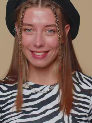 A young woman with fair skin and blue eyes looks directly at the camera with a slight smile. She wears a black beret, a silver earring, a zebra-striped top, and a silver chain necklace. Her long hair is parted and has braids, with red accents.