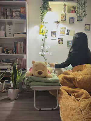 A cozy bedroom setting with a woman sitting on a bed covered in a soft blanket. A small table holds a Winnie the Pooh plush. White shelves filled with books and decorative items are also visible, giving the room a warm and inviting atmosphere.
