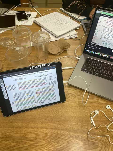 A top-down shot of a wooden table scattered with study materials. There's a laptop, a tablet displaying text from a document marked with annotations, three clear plastic cups, notebooks, a phone, and earphones. Stray wires and a paper bag are also visible. The lighting is natural, casting shadows, and the overall composition suggests a study environment.