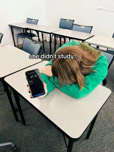 A person is sitting at a desk in a classroom setting, leaning their head down on the desk with long brown hair partially covering their face. They are wearing a green shirt or sweater. In their hand, they are holding a smartphone with a black screen that displays a percentage of 21% and other information. Several desks and chairs are visible in the background.