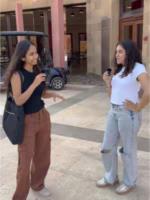 Two young women stand outside, facing each other and holding microphones. Buildings and a golf cart are in the background.