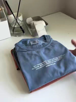 A stack of folded t-shirts in various colors is seen on a white surface. The top shirt is blue with white text, and the folded shirts beneath are red and gray. The setting is a white countertop with a few decorative items in the background like a glass diffuser and a small container with round stickers.
