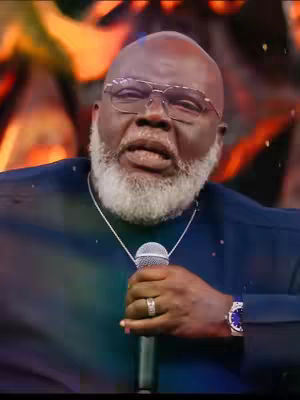 A medium shot features a bald, older Black man with a white beard and glasses. He is holding a microphone in his right hand, with his left hand near it. He is wearing a dark blue shirt and a silver necklace. Behind him, there appears to be a blurred background with hints of warm colors.