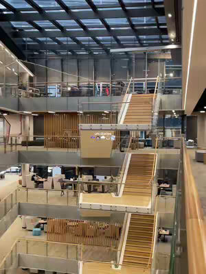 An expansive, multi-level university building interior. The structure features multiple floors with open walkways and large windows, creating a sense of spaciousness and natural light. Wooden accents and stairs add warmth to the modern design. Several people are visible, suggesting an active study environment.