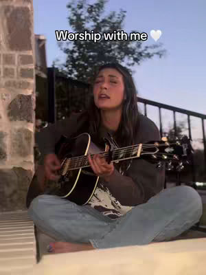 A woman sits cross-legged outdoors, playing an acoustic guitar. She has long, dark hair and is wearing a dark top and light blue jeans. The background shows a tree, a fence, and the evening sky. Text overlay: "Worship with me".